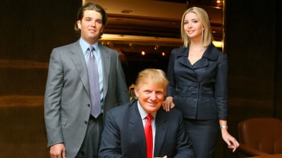 Donald Trump vid ett skrivbord omringad av sina barn Donald Trump Jr. och Ivanka Trump, alla ler och ser in i kameran.