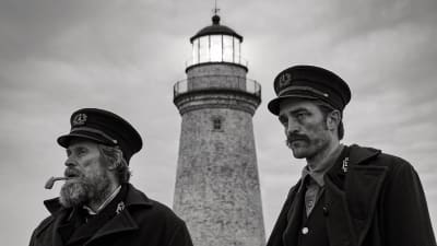En svartvit bild på Thomas Wake (Willem Dafoe) och Ephraim Winslow (Robert Pattison) står nedanför en fyr och tittar ut mot havet.