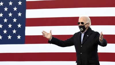 En man klädd i svart kappa och blå skjorta står med händerna utstäckta. Han har ett svart munskydd där det står Vote samt svarta solglasögon. Hans hår är vitt. I bakgrunden syns en enorm amerikansk flagga.