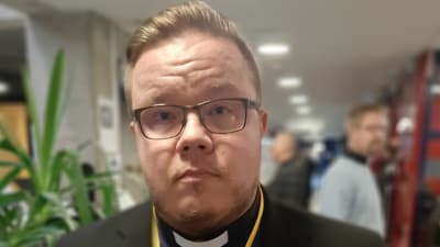 En man med ljust hår, mustasch, skägg och glasögon har en kavaj och blå skjorta med prästkrage.