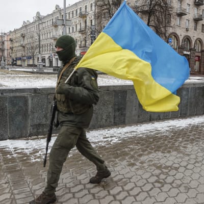 En soldat i militärutrustning går med ett vapen i ena handen och Ukrainas flagga i den andra. I bakgrunden en gammal byggnad.