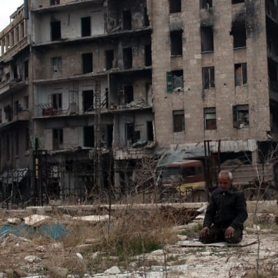 En man sitter på knä och ber i östra Aleppo, som är belägrat av rebeller. I bakgrunden övergivna byggnader skadade av striderna i staden.