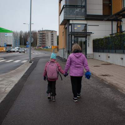 En kvinna och ett barn med ryggsäck fotograferade bakifrån där de går en en gångväg intill en gata.
