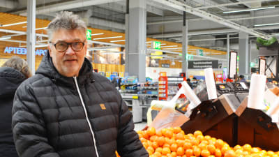 En medelålders man står i en matbutik bredvid en hög med mandariner. Han tittar in i kameran.