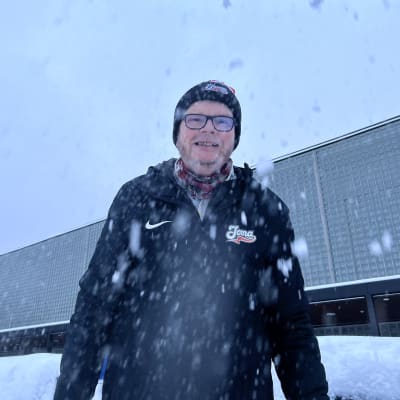 Mustaan talvitakkiin pukeutunut Ismo Björn hymyilee kameralle. Lunta sataa.