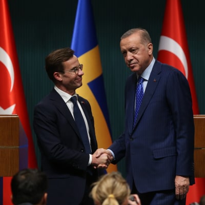 Sveriges statsminister Ulf Kristersson skakar hand med Turkiets president Recep Tayyip Erdoğan. Bakom de två männen syns en rad med flaggor, varannan turkisk och varannan svensk.