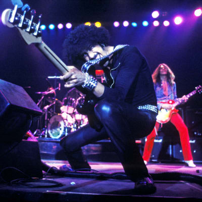 Phil Lynott osoittelee bassollaan lavalla, taustalla muita soittajia.