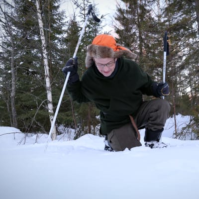 Oranssilakkinen Antti Suontama on kyykistynyt sauvoihin nojaten tutkimaan lumen pintaa.