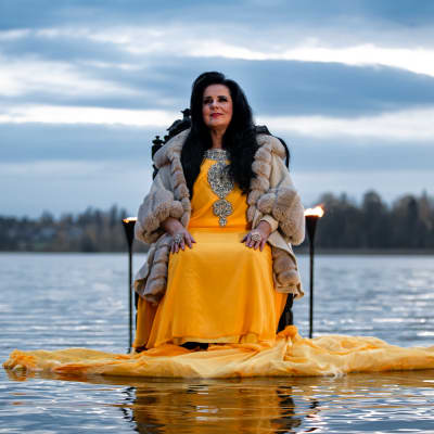 Kaija Koo istuu tuolilla veden keskellä Tuusulanjärven rannassa. Kaija Koolla on yllään keltainen mekko ja vaalea turkki.