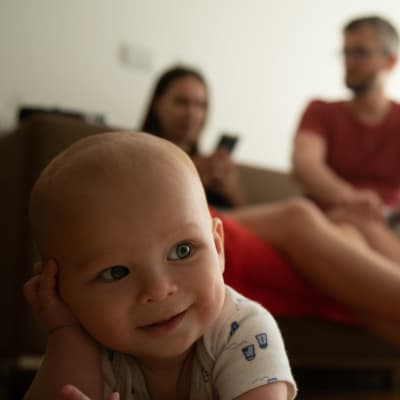 Kuvassa etualalla vauva katsoo kameraan, taikana sohvalla istuva pariskunta. 