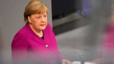 Angela Merkel talar framför ett talarpodium. Hennes reflektion syns på bildens högra sida.