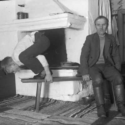 En man leker leken "ta sticka från långbänken", där han står på en bänk och hukar sig. Bilden är tagen 1936 i Strömfors, Kungsböle.