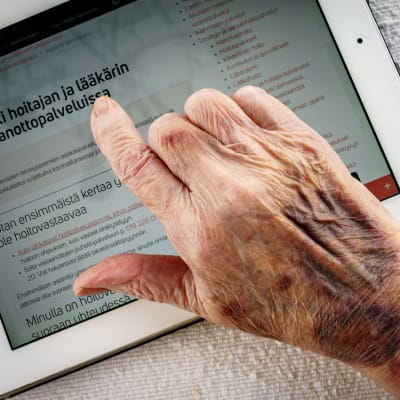 Vanhuksen ryppyinen ja mustelmainen käsi tablettitietokoneen ruudulla. Ruudulla lukee asiointi hoitajan ja lääkärin vastaanottopalveluissa.
