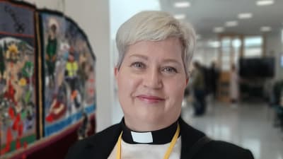 Heidi Juslin-Sandin, en dam med blont sidokammat hår, prästkrage, vit skjorta och svart kavaj, tittar rakt in i kameran.