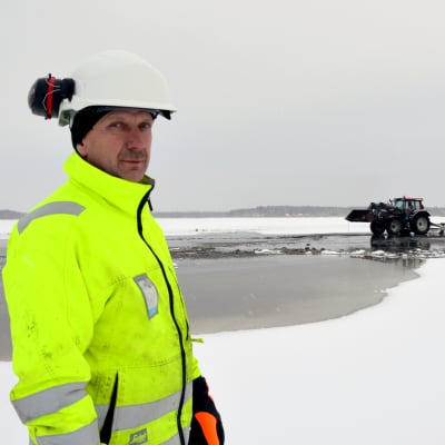 Yngve Sundström håller i trådarna för muddringen på Stadsfjärden i Vasa. Här står han på isen iförd en neongul arbetsmundering och hjälm. I bakgrunden ser man en traktor.