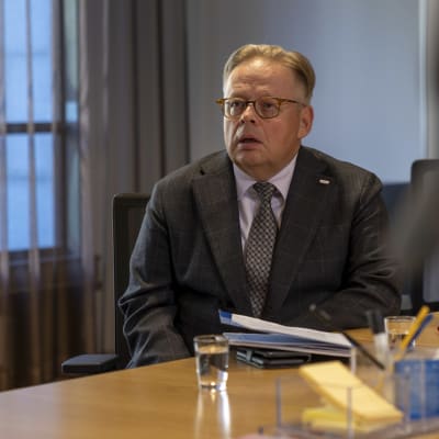 Helsingfors stads borgmästare Juhana Vartiainen sitter vid ett bord med en tunn mapp framför sig. I förgrunden skymtar journalister som deltar i den presskonferens där Vartiainen talar.