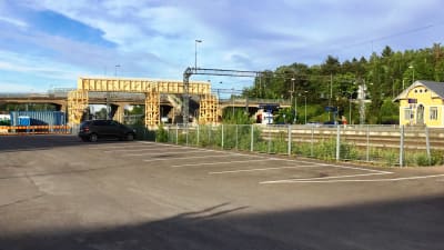 I förgrunden en tom asfalterad parkering. Längre borta syns en bro över en bangård samt en tillfälllig träbro. Karis järnvägsstations, en gul stockbyggnad, syns till höger.