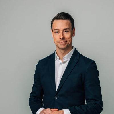 Mies (Jere Nurminen) seisoo tummassa puvuntakissa kädet ristissä ja hymyilee kameraan.