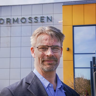 Aimo Latvala står utanför Stormossens byggnad. Han tittar in i kameran. Han är klädd i en grå kavaj och blå skjorta.