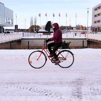 En kvinna cyklar förbi byggnader i betong och glas på en väg med ett tunt lager snö. Himlen är ljusblå och klar.