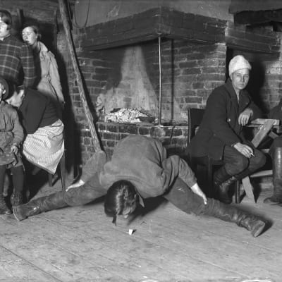 En man leker leken "ta spåna", där man stå så bredbent man kan och ta upp ett föremål från golvet utan att knäböja sig eller röra golvet med händerna. Bilden är tagen 1935 i Bromarf.