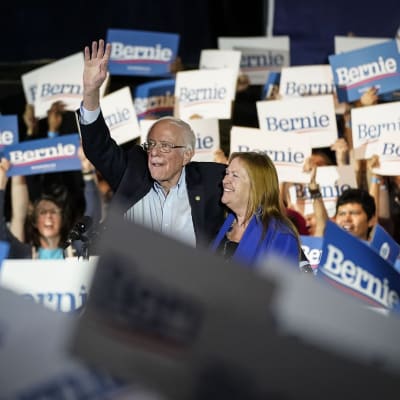 Bernie Sanders håller upp handen, hans fru ler bredvid. Runt honom glada anhängare men plakat det står Bernie på.