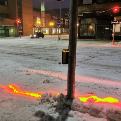 En gatukorsning, där en linje av rött lyser i snön samtidigt som det lyser rött för fotgängare.