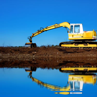 En grävmaskin gräver i torvjorden vid vattnet. Grävmaskinen speglas i vattenytan.