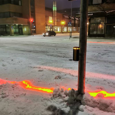 En gatukorsning, där en linje av rött lyser i snön samtidigt som det lyser rött för fotgängare.