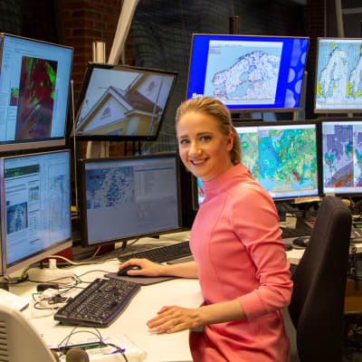 Meteorologi Anniina Valtonen hymyilee kameralle työpisteellään ja häntä ympäröi yhteensä 10 näyttöä, joilla on erilaisia sääkarttoja.