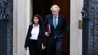 Munira Mirza (kavaj och skjorta) går ner för trappor framför Boris Johnson (kostym med slips).