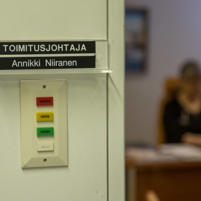 Työhuoneen ulkopuolella kyltti, jossa lukee "toimitusjohtaja Annikki Niiranen". Epätarkkana taustalla näkyy huoneessa henkilö työpöydän ääressä.