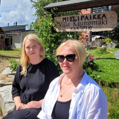 Kaksi naista istuu puutarhassa, taustalla kyltti jossa lukee Mielipaikka Villa Kaunismäki Kakspyy.