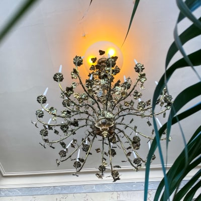 En takkrona i guld med ljus i. Fotograferad från bakom en grönväxt så att några blad synns i förgrunden.