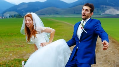 kvinna i brudklänning lutar sig framåt och ler, man i blå kostym ser förskräckt ur