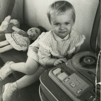 Kaksivuotias Sara Paavolainen nojaa isänsä Grundig magnetofoniin. Taustalla nukke ja nalle.
