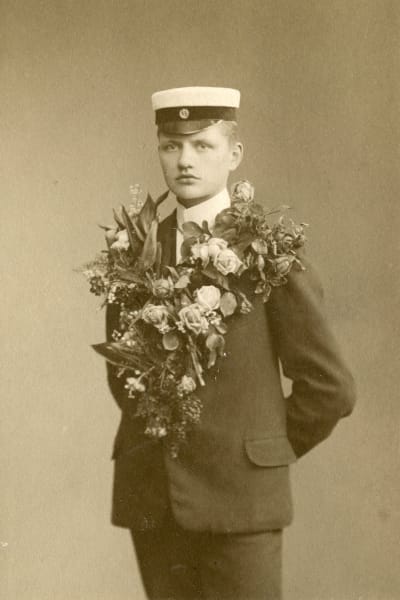 Carl Nymans studentfoto där han står med allvarlig min och en enorm blomsterklass runt halsen.