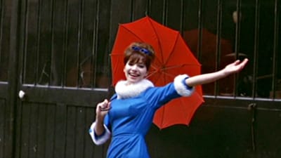 Anna Karina hymyilee kameralle punainen sateenvarjo kädessä. Kuva elokuvasta Nainen on aina nainen.
