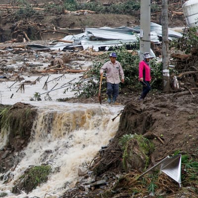 Två personer går vid ett område som förstörts av jordskred och översvämning. Det forsar vatten överallt.