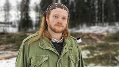 En person tittar in i kameran. Hen ser nyfiken men allvarsam ut. I bakgrunden ett kalhygge.