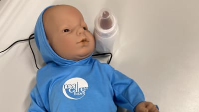 En baby-docka iklädd en blå tröja och blåa byxor. En sladd löper från dockan ur bilden. Bredvid dockan en nappflaska.
