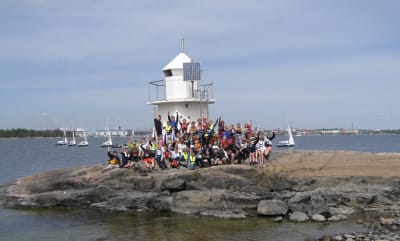 Joukko nuoria purjehtijoita on kavunnut maihin pienelle luodolle jossa on pieni majakka. Taustalla purjejollia ja Helsinki mereltä nähtynä.