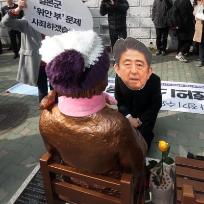 En man iförd en mask, som föreställer Japans premiärminister Shinzo Abe, knäböjer framför statyn i Busan 4.1.2017