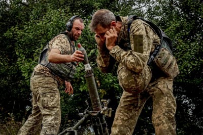 Två camouflageklädda soldater gör sig redo att avfyra en granatkastare. Den ena lägger en granat i röret och den andra har fingrarna i öronen.