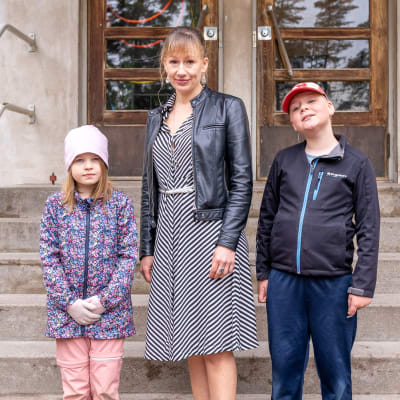 Två elever och en lärare står vid en stentrappa till en gammal skolbyggnad från 1950-talet. Den ena eleven har axellångt hår och en rosa möss och blommig rock och den andra röd keps och svart jacka. Läraren bär randig klänning och en svart läderjacka.