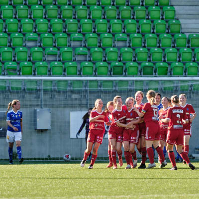 En klunga unga kvinnor i röd spelarklädsel gratulerar varandra glatt efter match. I bakgrunden ett fotbollsmål.