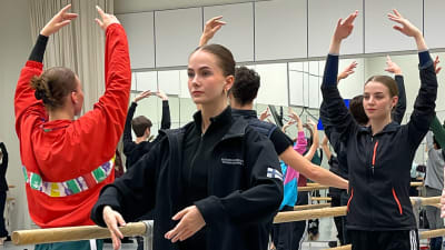 Balettitanssijoita harjoitussalissa tekemässä tankoliikkeitä varpaillaan seisten ja kädet kohotettuna ylös. Kasvoilla keskittynyt ilme.
