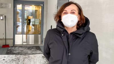 En kvinna i vinterjacka och munskydd står utanför ingången till ett servicehem.