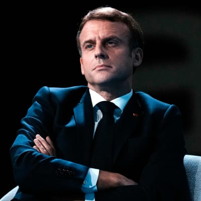 Emmanuel Macron sitter på en stol med armarna i kors. Han ser allvarlig ut medan han tittar åt sidan. Han har på sig en mörk kostym.