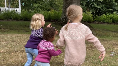 Tre barn springer på en gräsmatta iklädda handstickade strömsöbarntröjor.